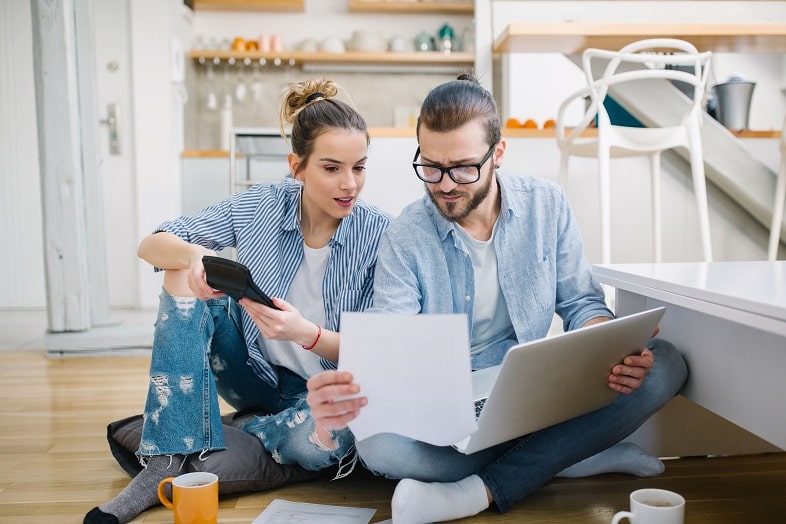 Mulher segurando uma calculadora e olhando um papel que está sendo segurado por um homem com um notebook no colo